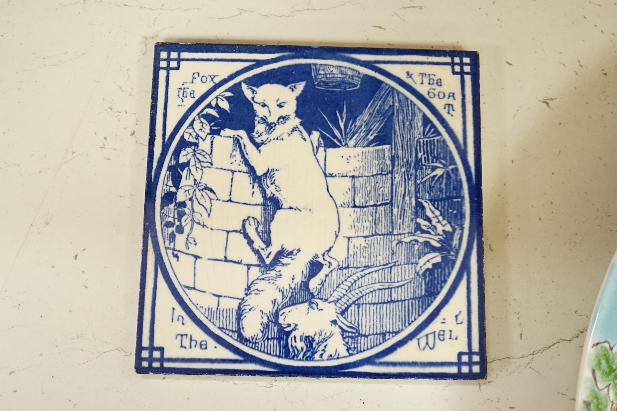 Two Minton pottery Arts and Crafts fireplace tiles, printed in blue and white, with scenes from Aesops fables - ‘’The Fox and the Goat in the Well’’ and ‘’The Hare and the Tortoise’’, together with a Poole pottery dish.